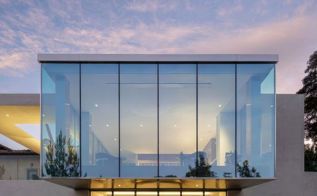 Contemporary glass-fronted building extension photographed from outside at dusk. A broad rectangular volume is enclosed by floor-to-ceiling blue-tinted glazing with slim dark mullions and a flat projecting roof slab. Warm interior lighting glows through the lower level, while adjacent solid rendered walls and a curved pale-yellow architectural element are visible at the sides.