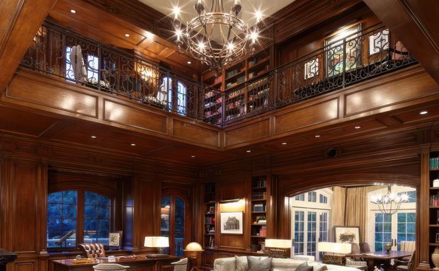 Opulent double-height library or living space with extensive dark timber wall panelling, coffered timber ceiling, built-in bookshelves, ornate wrought-iron balcony railings, arched windows and doors, and a prominent multi-arm chandelier. A mezzanine gallery overlooks the furnished lower level, creating a grand traditional interior with warm ambient lighting.