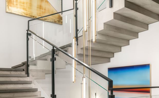 A contemporary interior stairway with two flights of floating concrete stairs, slim dark metal handrails, and clear glass balustrades. Vertical pendant lights hang in the stair void, while abstract artwork adds colour to the otherwise light, minimalist setting.