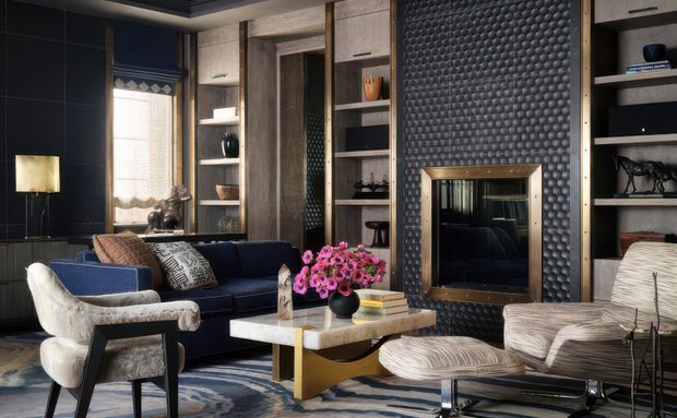 A sophisticated, dark-toned living room with a navy sectional sofa, sculptural upholstered lounge chairs, a marble-topped coffee table with a gold-toned base, and built-in shelving. The walls feature dark panelling and a dramatic textured geometric fireplace surround with a brass-framed firebox. Layered textiles, decorative objects, and floral arrangements add warmth and refinement.