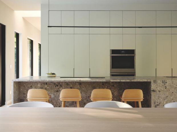 A bright contemporary kitchen viewed across a timber dining table. The space features a large stone island with an integrated dark stone waterfall-style front, four timber bar stools, pale full-height flat-panel cabinetry, and a built-in black oven. Tall narrow windows and a skylight-like opening bring in natural light, creating a warm, calm, minimalist interior.