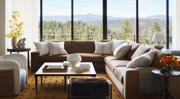 A warm, refined living room with a large L-shaped brown upholstered sectional, white accent cushions, a low rectangular coffee table, and a textured ottoman. Floor-to-ceiling glazing frames expansive forested mountain views, while neutral drapery, a patterned rug, side tables, lamps, and floral arrangements complete the inviting interior.