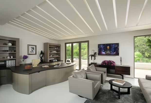 A refined open-plan living room with an integrated home bar or kitchenette at left and a lounge area at right. The room features a curved dark benchtop with grey fixed cabinetry, built-in shelving, a wall-mounted television, contemporary armchairs and sofa, a dark rug, and a low round coffee table. Floor-to-ceiling glazing and glazed doors open toward a leafy outdoor setting. White linear ceiling battens create a strong architectural feature across the room.