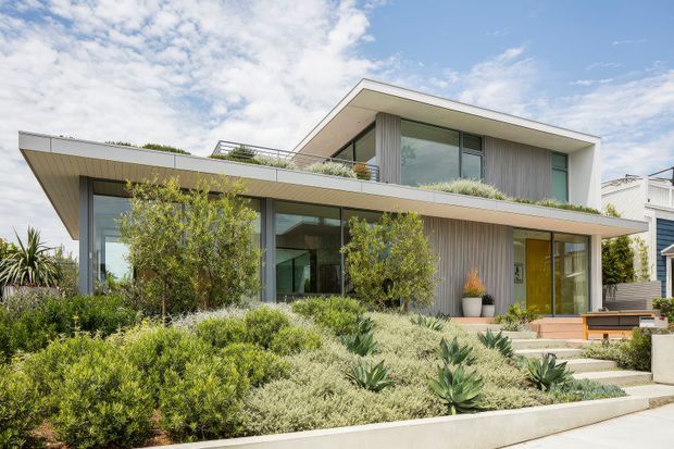 Contemporary two-storey residence viewed from the landscaped front garden, featuring broad horizontal roof overhangs, extensive glazing, pale rendered walls, vertical timber-look cladding, and a warm timber-toned entry door. The house is integrated into a drought-tolerant garden with shrubs and sculptural succulents, with a rooftop terrace or planted upper-level outdoor area visible behind the parapet.