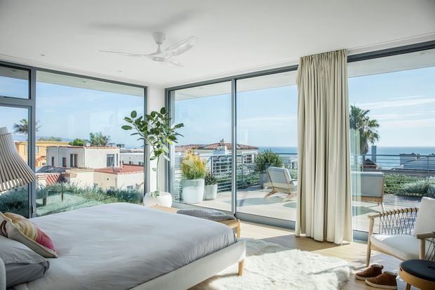 Bright, airy bedroom with a large upholstered bed positioned beside floor-to-ceiling glazing. The room opens onto a balcony or terrace with outdoor lounge chairs and expansive coastal views. Neutral white and cream finishes, light timber accents, indoor plants, sheer curtains, and a ceiling fan create a relaxed contemporary coastal atmosphere.