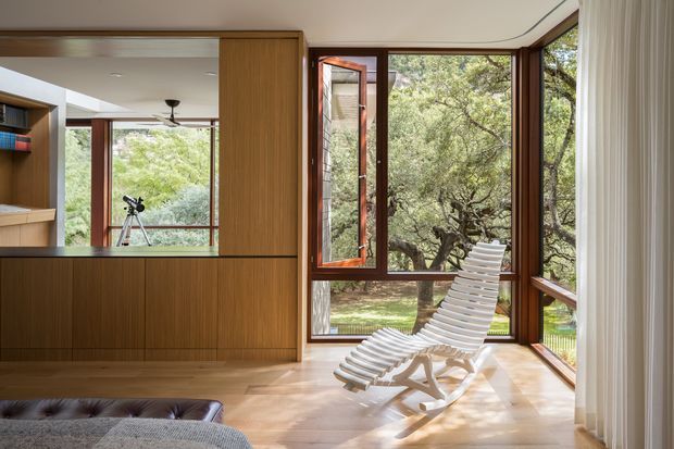 A calm, light-filled modern interior with floor-to-ceiling timber-framed glazing overlooking mature trees. Built-in warm timber joinery with open shelving and a concealed doorway divides the room, while a white sculptural lounge chair sits on pale timber flooring beside the windows. A partial upholstered seating area is visible in the foreground, and the glazing includes an open casement window.