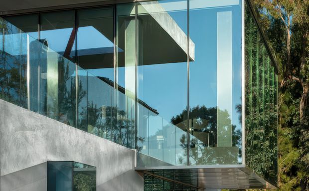 Exterior architectural photograph of a contemporary building set among trees, featuring extensive floor-to-ceiling glass walls, a projecting glazed volume, exposed concrete walls and a cantilevered form. Reflections reveal the surrounding vegetation and blue sky; no interior room is clearly identifiable.