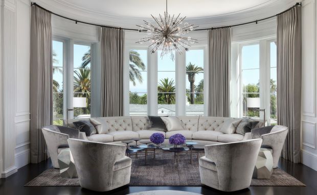 Luxurious, symmetrical living room with a large curved tufted sectional sofa, sculptural lounge chairs, nested coffee tables, and a dramatic starburst chandelier. Tall bay-style windows with layered grey drapery overlook palm trees and a bright coastal scene. White decorative wall mouldings, dark timber flooring, and a neutral grey palette create a refined, glamorous interior.