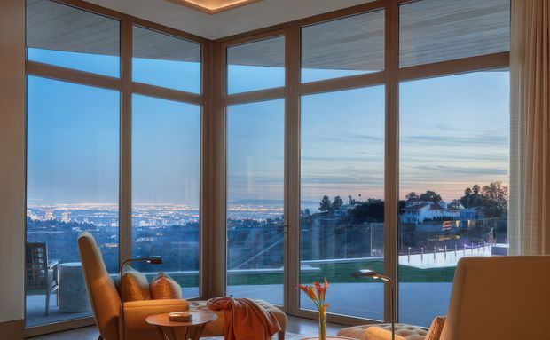 A refined living room or sunroom with expansive floor-to-ceiling corner glazing overlooking a suburban cityscape at dusk. Warm timber-framed windows, a recessed ceiling detail with concealed lighting, upholstered lounge seating, and a small glass coffee table create a calm, luxurious atmosphere. A terrace, pool, and landscaped area are visible outside.