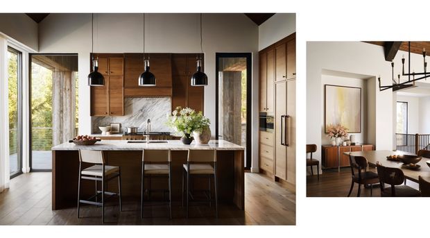A refined open-plan kitchen and dining area. The kitchen features a dark timber island with seating for four, warm timber cabinetry, a pale veined stone benchtop and splashback, black pendant lights, and large glazed doors. A connected dining space is visible at right with a timber dining table, dark chairs, a sideboard, artwork, and a linear chandelier. The overall scheme is warm, contemporary, and carefully detailed.