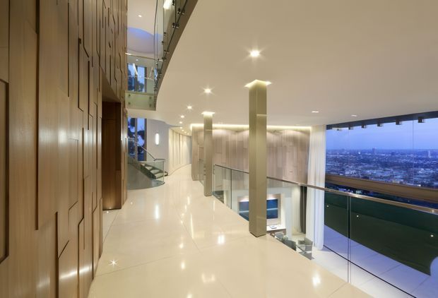 A refined, multi-level interior landing or hallway with glossy pale flooring, tall integrated timber wall panelling, slender square columns, recessed ceiling lights, glass balustrades, and a curved stair visible at the rear. Full-height glazing opens to an expansive city view, while the void below reveals another level of the home. The space has a polished, luxurious contemporary character.