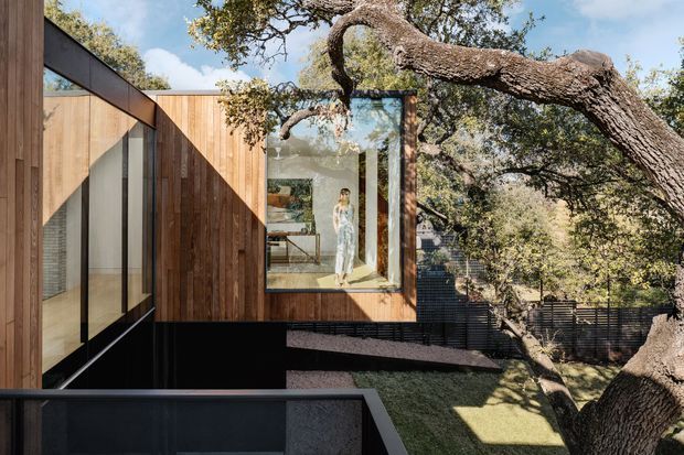 Exterior view of a contemporary timber-clad residence integrated among mature trees on a sloping site. Dark metal framing and large floor-to-ceiling glazing create a strong contrast with the warm vertical wood cladding, while an interior living space is visible through the central window.