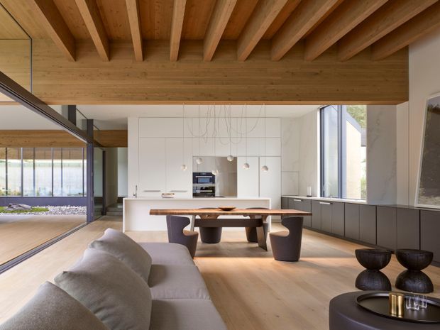 Open-plan contemporary living, dining and kitchen interior with extensive warm timber detailing. A white kitchen with integrated appliances and island sits behind a dark timber dining table, while a large pale sectional sofa occupies the foreground. The room features a dramatic exposed timber beam-and-joist ceiling, full-height glazing, dark built-in cabinetry, pendant lighting and a calm neutral palette.