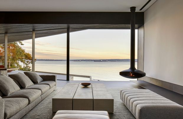 Contemporary waterfront living room with expansive floor-to-ceiling glazing overlooking calm water. The space features a large grey sectional sofa, matching upholstered bench seating, a low rectangular coffee table, dark structural columns and ceiling detailing, and a suspended black fireplace. The restrained neutral palette and open outlook create a refined, serene atmosphere.