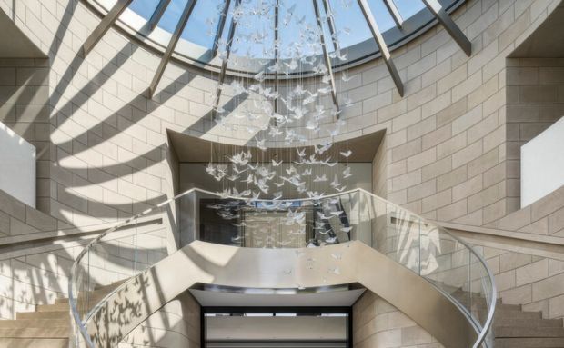 A dramatic multi-level interior atrium with curved staircases and transparent glass balustrades, enclosed by pale masonry block walls. A large glazed skylight floods the space with daylight, while a suspended installation of delicate white bird-like forms hangs through the central void.