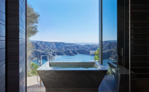 A dramatic bathroom with a freestanding dark stone bathtub positioned before full-height glazing, overlooking an infinity pool and a mountainous turquoise lake landscape. Dark vertical wall cladding, reflective glazing, and strong indoor-outdoor visual continuity create a luxurious, contemporary retreat.
