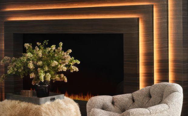 A warm, dramatic living room focal wall featuring a large black fireplace opening framed by layered dark timber wall panelling and concealed amber lighting. A sculptural textured armchair sits in the foreground beside a floral arrangement and a soft sheepskin-covered surface.