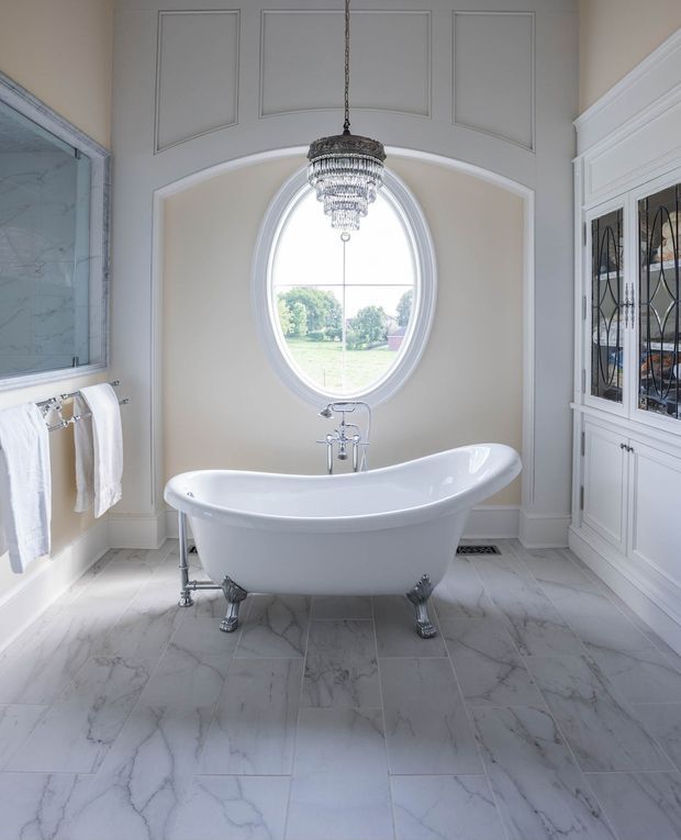 Bright, elegant traditional bathroom featuring a freestanding white clawfoot bathtub beneath an oval window. The room has pale marble-look tiled flooring, white built-in cabinetry with mirrored leaded-glass doors, a towel rail, decorative wall panel mouldings, and a crystal pendant chandelier. The palette is predominantly white, cream, and light grey with refined classic detailing.
