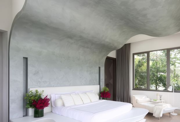 A contemporary bedroom with a large low-profile white bed positioned against a textured grey feature wall. The ceiling and wall form a sculptural, flowing plastered surface that curves organically around the room. Floor-to-ceiling dark curtains frame a broad window overlooking trees, while white upholstered seating and side tables create a soft, refined interior. Floral arrangements add small red and green accents.