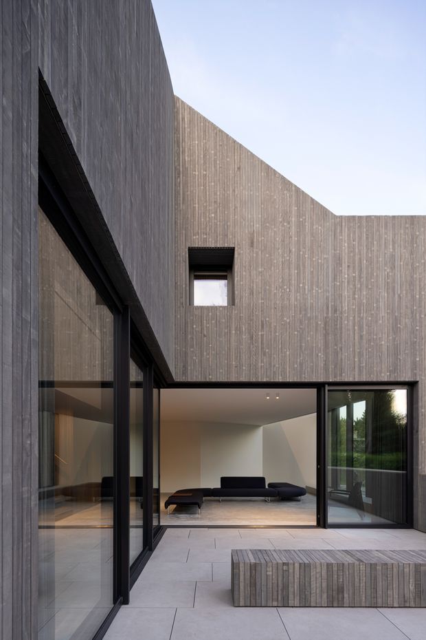 A minimalist contemporary courtyard exterior framed by tall vertical timber-clad walls. Large black-framed glazed openings connect the paved courtyard to a sparsely furnished living room, with a low dark sectional sofa visible inside. The courtyard includes a substantial built-in timber-clad bench or planter-like element, while the architecture features strong geometric planes, concealed rooflines, and carefully controlled openings.
