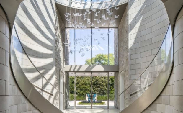 A bright, double-height architectural entry foyer with curved pale stone or concrete-block walls, expansive floor-to-ceiling glazing, and a dramatic suspended flock of bird sculptures. Strong sunlight creates patterned shadows across the walls, while a glazed entrance opens toward landscaped grounds.