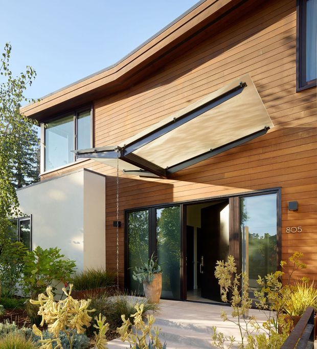 Exterior view of a contemporary timber-clad residence with a low-pitched roof and dark-framed glazing. The facade features a large outward-opening awning window above the entry, full-height glass doors, horizontal wood siding, and a landscaped approach with drought-tolerant planting.