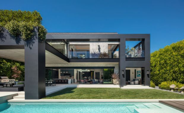 Exterior view of a large contemporary two-storey residence with a dark, rectilinear facade, extensive floor-to-ceiling glazing, covered outdoor living areas, landscaped lawn, and a swimming pool in the foreground. The open-plan interiors and upper-level voids are visible through the glazing.