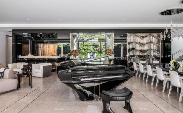 A spacious, highly refined open-plan living and dining area centered around a glossy black grand piano. The room features large pale stone-look floor tiles, dark built-in cabinetry and wall panels, expansive glazing, a long white dining table with white upholstered chairs, contemporary seating, sculptural lighting, and a dramatic crystal chandelier. The overall aesthetic is luxurious and contemporary with strong black-and-white contrast.