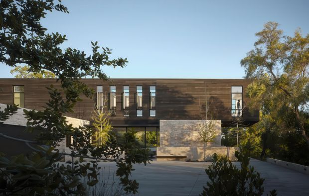 Exterior view of a contemporary two-storey residence set among mature landscaping. The building has a long, low horizontal form with dark timber-look cladding, narrow vertical windows, extensive ground-level glazing, and a projecting upper floor. A pale stone or concrete feature wall anchors the centre, while a broad paved terrace and surrounding trees frame the foreground.