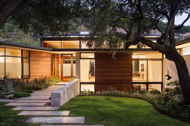 Single-storey modern residence viewed from a landscaped front garden, with broad low-pitched roof planes, extensive floor-to-ceiling glazing, warm horizontal timber wall cladding, pale masonry walls, and a stepped concrete walkway leading to the entry. Mature trees frame the house and partially screen the facade.