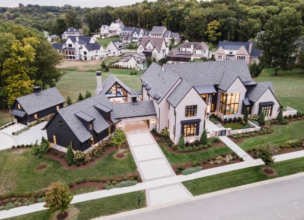 Aerial view of a large, multi-building residential property in a landscaped suburban development. The main house features steep gabled roofs with dark grey shingles, light rendered walls, black-clad ancillary buildings, extensive glazing, paved pathways, planted garden beds, and a broad driveway. Similar large homes are visible across the wooded site.