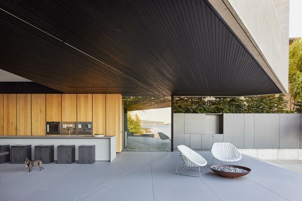 Contemporary covered outdoor living area with a dramatic cantilevered dark timber-lined soffit, integrated timber kitchen cabinetry and island, and a minimalist seating zone. Pale concrete paving, a light rendered privacy wall, sculptural lounge chairs, and a low fire bowl create a refined indoor-outdoor entertaining space. Water and distant hills are visible beyond the glazing.