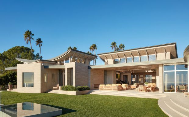 A large contemporary single-storey residence viewed from the landscaped front lawn, featuring broad glazing, pale masonry or rendered walls, warm timber-toned cladding, deep roof overhangs, and distinctive curved and angled roof forms. A covered outdoor entertaining area opens toward the lawn and reflective water feature, with palm trees and clear blue sky in the background.