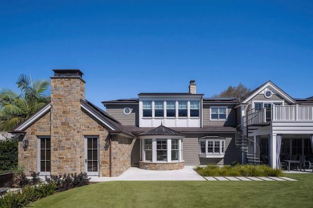 Exterior view of a large multi-wing residential house surrounding a landscaped lawn and paved courtyard. The home combines dark horizontal cladding, extensive stone veneer, pitched gable roofs, dormer-like upper sections, white-trimmed windows, chimneys, and a raised deck with an exterior spiral staircase. Bright clear daylight emphasizes the formal, refined coastal-residential character.