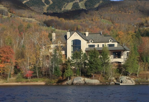 Exterior view of a large multi-storey house set directly along a wooded lakeshore, with a steep dark gabled roof, pale masonry or stucco walls, prominent tall arched window, dormer windows, covered balcony or terrace, and multiple chimneys. Autumn forested hills rise behind the property, while exposed rock and shoreline vegetation sit between the house and the water.
