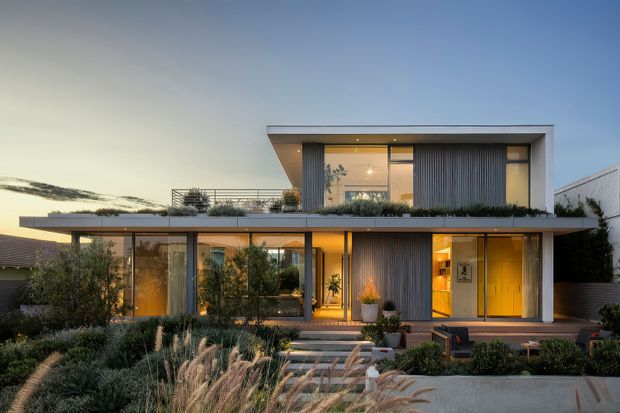 Twilight exterior view of a contemporary two-storey residence with a crisp flat-roofed form, extensive glazing, dark vertical ribbed cladding, warm interior lighting, rooftop terrace-style planting, and a landscaped front garden with paved entry steps.