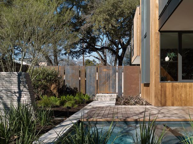 A contemporary residential exterior viewed from a landscaped garden, featuring warm vertical timber cladding, dark-framed glazing, a weathered metal privacy fence, a paved timber-toned terrace, and a narrow blue swimming pool bordered by natural planting. Mature trees and layered greenery create a private courtyard-like setting.
