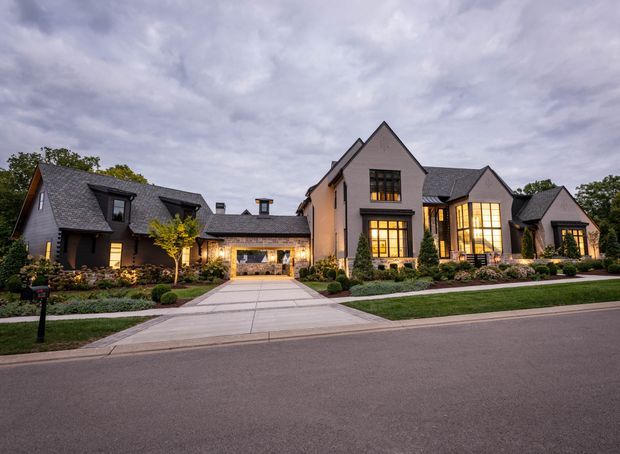 A large, multi-wing suburban residential house photographed from across the street at dusk. The home has steep gabled roof forms, dark grey shingle roofing, light rendered masonry walls, dark-framed windows with warm interior lighting, landscaped garden beds, and a broad concrete driveway leading to a central recessed garage or entry bay. The composition is an exterior architectural elevation with several connected gabled volumes.