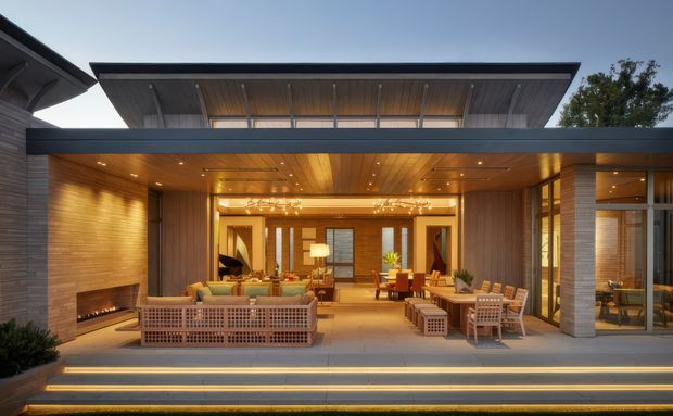 A wide exterior view of a sophisticated contemporary residence with a covered outdoor entertaining area opening into a warmly lit living and dining space. The home features strong horizontal roof planes, extensive glazing, timber-lined ceilings and walls, integrated recessed lighting, masonry walls, broad steps, and built-in outdoor seating and dining furniture.