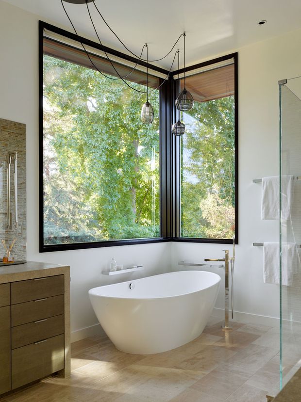 Bright contemporary bathroom with a freestanding white oval bathtub positioned in front of large corner windows overlooking leafy trees. The room includes a floating timber-toned vanity with integrated basin, a glass-enclosed shower, wall-mounted towel rails, pale stone-look floor tiles, dark window frames, and sculptural pendant lights with exposed cords.