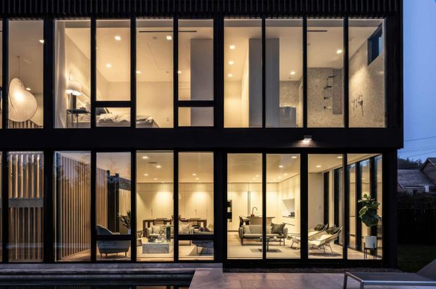 Dusk exterior view of a contemporary multi-storey residence with a dark framed glazed facade. Floor-to-ceiling windows reveal a warmly lit living area, dining space, bedroom, and tiled bathroom inside. Vertical timber screening and a paved outdoor area frame the house.