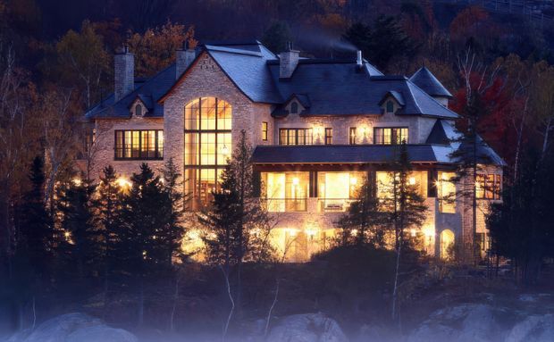 A large multi-storey stone-clad residence photographed at dusk in a wooded, mountainous setting. The house has a complex pitched and gabled roofline, multiple chimneys, prominent warm-lit windows including a tall central glazed opening, and a projecting lower-level terrace or enclosed porch. Dense trees partially screen the building.
