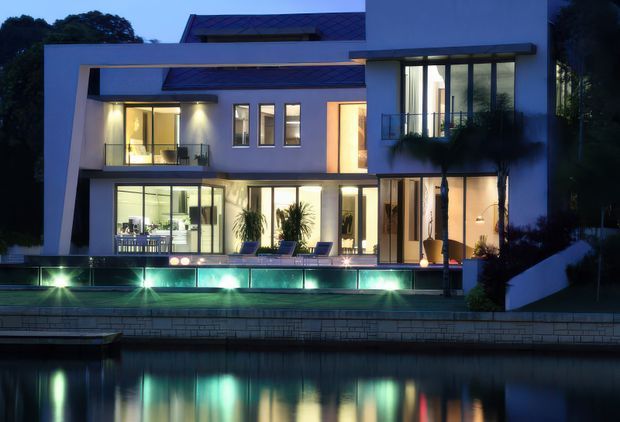 Twilight exterior view of a contemporary multi-level residence with crisp white rendered walls, extensive floor-to-ceiling glazing, projecting rectangular roof and balcony forms, and a long illuminated swimming pool terrace. The house is reflected in a dark body of water in the foreground, with landscaped planting around the facade.