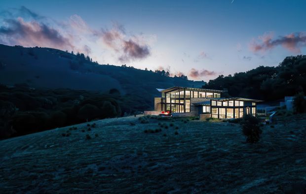 A modern, low-profile house glowing with warm interior light at dusk, set into a sloping mountainous or rural landscape. The building features extensive floor-to-ceiling glazing, crisp rectangular volumes, dark structural framing, and a broad shallow roofline. The surrounding terrain is dark and densely vegetated, with a blue twilight sky and scattered clouds.