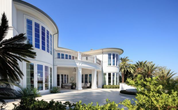 A grand white multi-storey residence with curved wings, tall divided glazing, classical columns, ornate balustrades, and broad paved courtyard or driveway. Tropical palms and lush planting frame the bright coastal-looking exterior.
