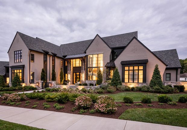 Large multi-gabled suburban residence photographed from the landscaped front yard at dusk. The façade combines pale rendered masonry with dark brick accents, steep charcoal shingle roofs, tall black-framed windows, and a prominent two-storey glazed entry volume. Formal planting beds, clipped shrubs, stone paths, and a concrete footpath frame the front elevation.