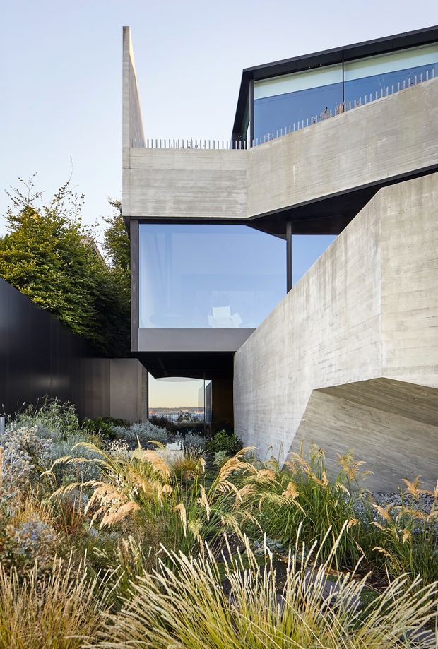 Exterior view of a striking contemporary concrete residence with layered cantilevered forms, dark metal framing, expansive glazing, and a sculptural angular stair or retaining-wall structure. The building is set within a lush ornamental grass and drought-tolerant garden, with glimpses of a distant urban or coastal landscape through the lower opening.