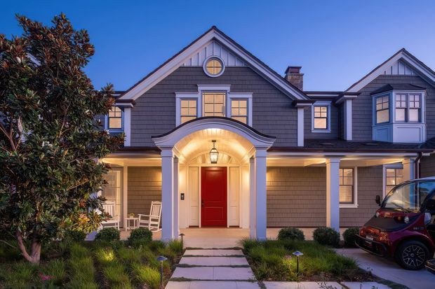A twilight front exterior view of a large, symmetrical shingle-clad home with multiple gabled roof sections, dormer-like upper windows, a covered entry porch, and a prominent arched portico framing a bright red front door. Warm interior and porch lighting contrast with the deep blue evening sky. A paved walkway crosses a landscaped front garden toward the entrance, with a parked vehicle partially visible at the right edge.