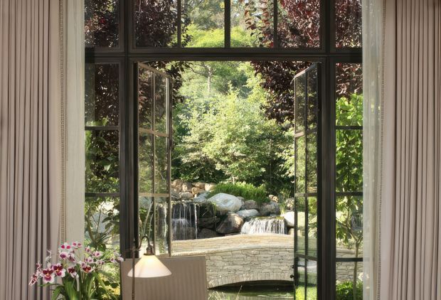 A refined interior view through tall, dark-framed multi-pane French doors and transom glazing, opening onto a lush landscaped garden with a stone-lined stream and small cascading waterfall. Sheer curtains frame the opening, while a table lamp and flowering orchid are visible in the foreground. The image emphasizes the strong indoor–outdoor connection and garden outlook.