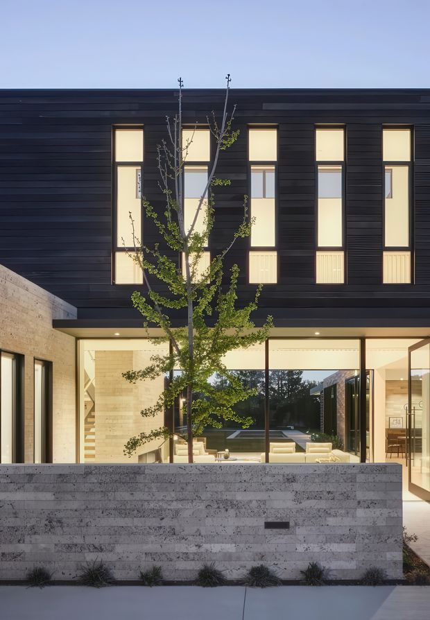 Contemporary two-storey residential facade viewed from the street at dusk. The upper level is clad in dark horizontal boards and punctuated by tall, narrow illuminated windows. The lower level has extensive floor-to-ceiling glazing opening toward a landscaped courtyard, with a pale masonry side wall and low stone boundary wall in the foreground. A slender deciduous tree partially screens the facade.